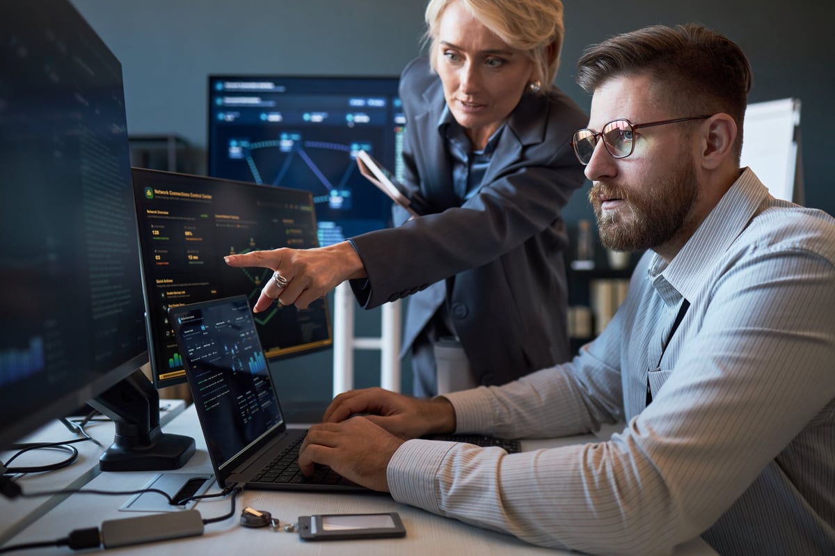 Caucasian businesswoman pointing at cybersecurity dashboard while Caucasian businessman analyzing data on laptop and monitors. Ideal for IT teamwork, network security, risk management